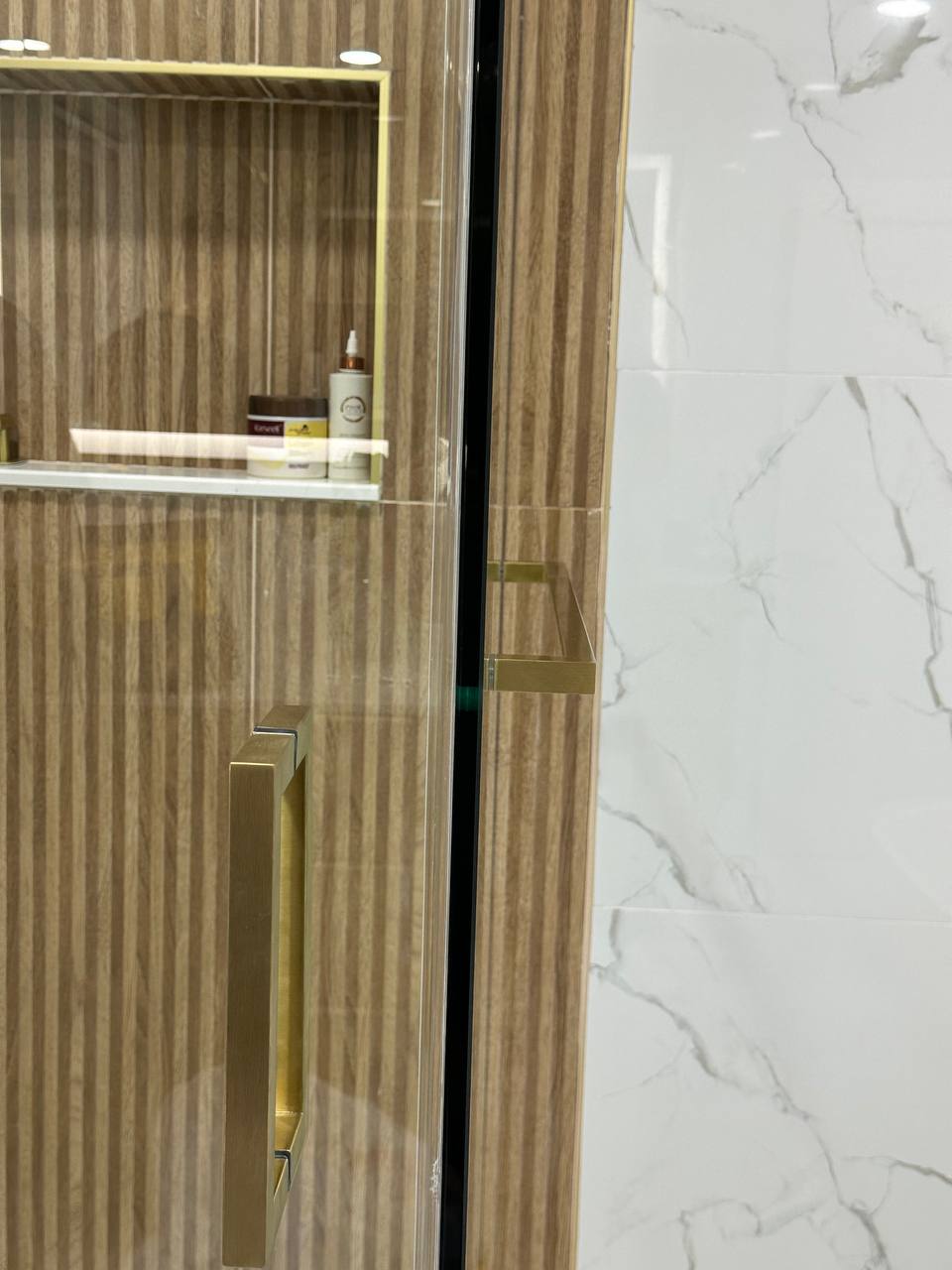 Close view of a brushed brass shower door pull on clear glass with warm vertical tile behind it.