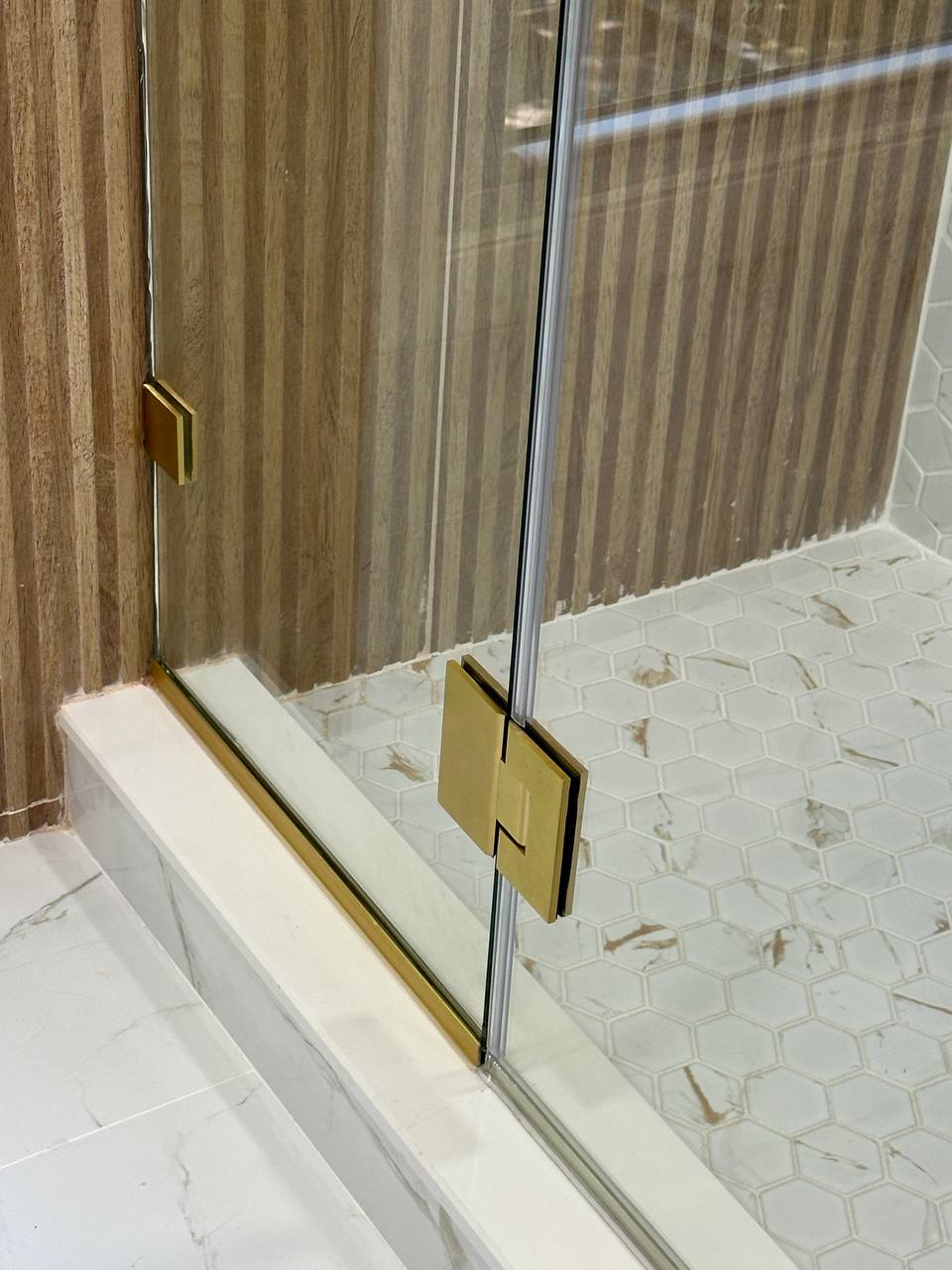 Lower brushed brass shower door hinge, channel, and clear glass edge above a white hex mosaic shower floor.