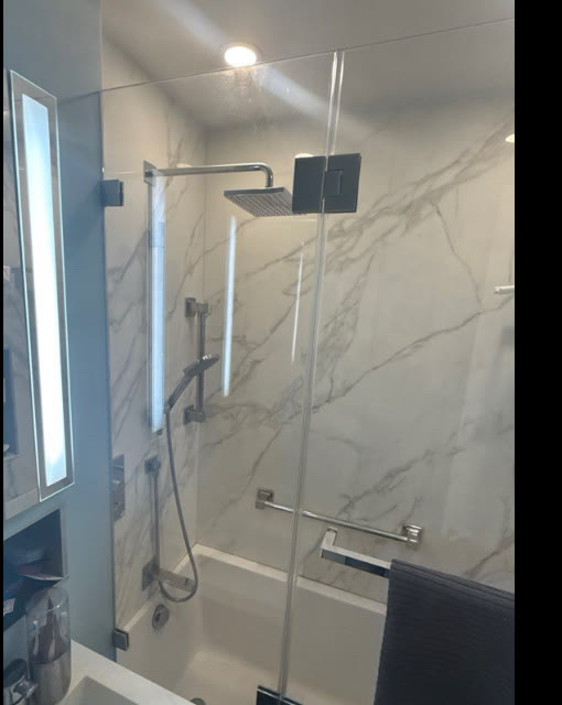 Frameless tub enclosure with chrome hardware and rain shower head against white marble tile, remediated NYC project