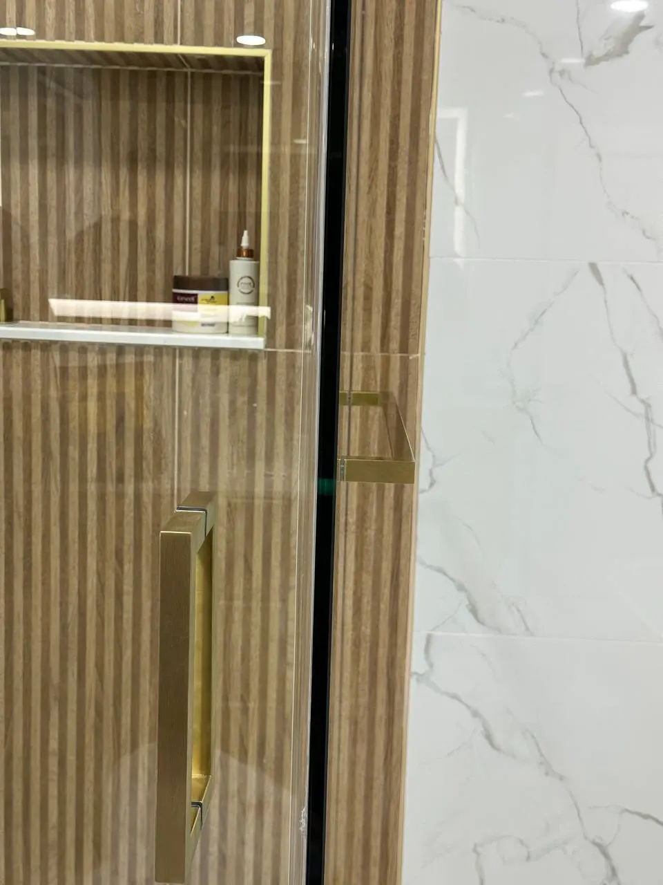 Close view of a brushed brass shower door pull on clear glass with warm vertical tile behind it.
