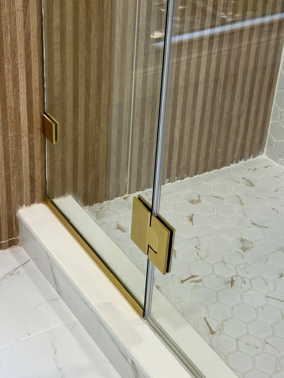 Lower brushed brass shower door hinge, channel, and clear glass edge above a white hex mosaic shower floor.
