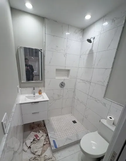 Open shower area before frameless glass installation, white marble tile throughout, floating vanity and built-in niche visible